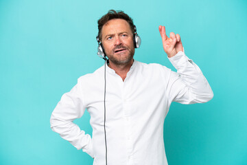 Telemarketer caucasian man working with a headset isolated on blue background with fingers crossing...