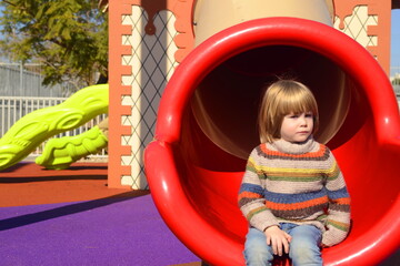 A sad boy sits on a slide in a playground. serious child in sweater, boy bad mood, boredom