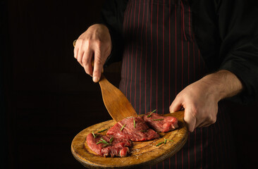 The chef turns over raw beef steaks with a wooden spatula before barbecue. Preparation for roasting meat with rosemary and spices. Black space for hotel recipe or menu.