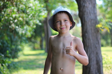 A smiling white schoolboy without clothes in the summer holds a thumbs up against the backdrop of a...