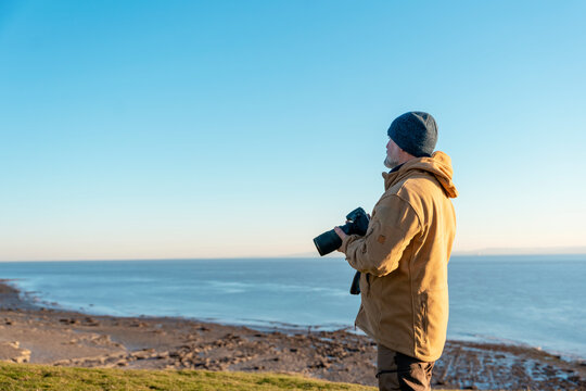Bearded Man reaching the destination  at sunset on cool day and taking photos of amazing seaside landscape in Wales. Travel Lifestyle concept.