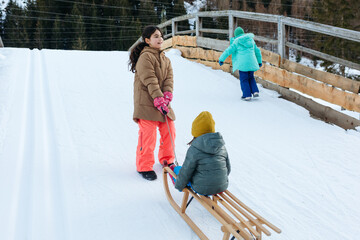 three children going up to snow slope with small sister being pulled by bigger one on wooden sled