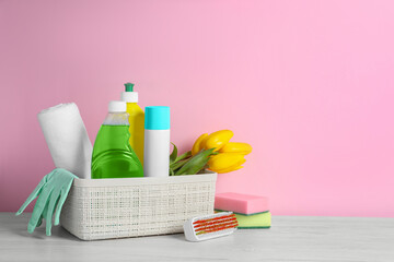 Fototapeta na wymiar Plastic basket with different cleaning supplies and beautiful spring flowers on white wooden table against light pink background. Space for text
