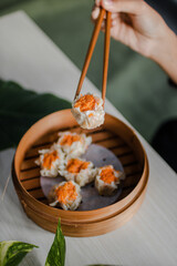 Female hand taking Chinese Dimsum with chopsticks in traditional bamboo basket on wooden table and green leaves background