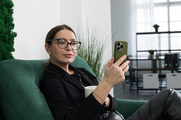 Serious young woman surrounded by plants sits in a chair and looks at the phone with a cup in hand.