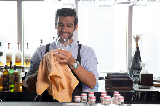 Young caucasian man is bartender cleaning and wipe glass with professional at counter, portrait handsome male preparation glass or wineglass, barman standing with confident at restaurant luxury.