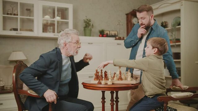 The grandson puts a checkmate to the grandfather. A boy wins chess from a surprised elderly man. Three generations of men from the same family play chess at home. A family hobby.
