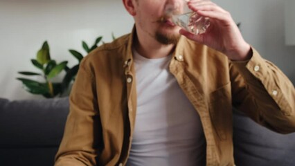 Selective focus of young bearded man taking medicine pill with water sitting on cozy sofa at home. Guy takes medication prescribed by his physician. Medicine, health care, pharmacy and people concept