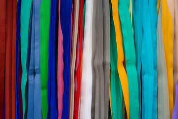 Many colorful zippers hang vertically next to each other from top to bottom in a tailor's shop. Different colors in a row next to each other