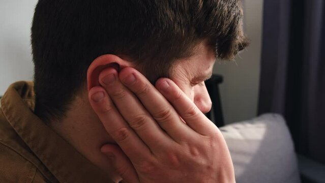 Closeup up side view of sick young caucasian male having ear pain touching painful head sitting on couch at home. Unhealthy man 30s suffering from painful otitis. Health care and tinnitus concept