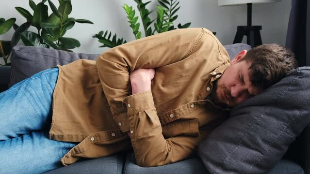 Unhealthy young man lying on sofa at home, suddenly feeling strong stomach ache, gastritis problem. Sad male squeezing belly with hands because of abdominal pain. Guy suffering from stomach ache