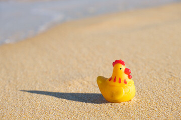 Fototapeta na wymiar Funny rubber toy yellow cockerel on a sandy beach in the foam of sea waves. Concept travel and vacation