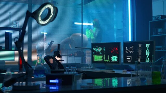 Modern medical research laboratory. Empty workplace of a scientist or researcher with computers, microscope, test tubes, flasks. Scientists in white protective overalls are working in the background.