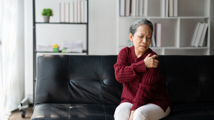 Senior Asian woman rubbing her shoulder, touch her shoulder to release the pain while sitting on the black sofa.