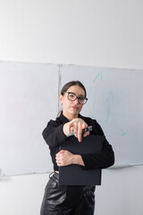 Professional woman in glasses and black suit holding a folder in front of a white marker board, explaining to colleagues in the office