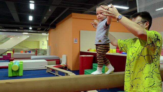 Baby boy is engaged with his dad on the gymnastic balance beam in a gym. FOOT USA MAIN