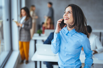 Happy young businesswoman smiling at the camera while holding a smartphone. Cheerful young businesswoman with her colleagues in the background. Just one call brought in a big deal