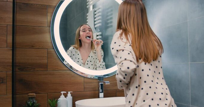 Funny Girl Is Singing And Dancing In Bathroom Wearing Pajamas Having Fun Alone At Home. Woman In Front Of The Mirror Holding A Toothbrush Dances And Sings.