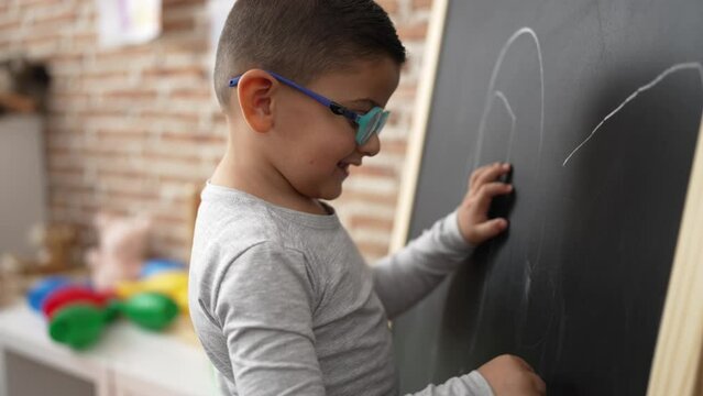 Adorable hispanic toddler student smiling confident drawing on blackboard at kindergarten