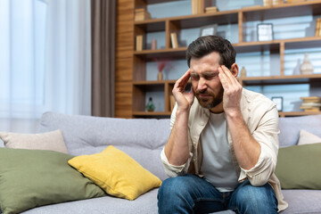 Fototapeta premium Headache of mature single man sitting on sofa alone at home and holding head with hands in living room.