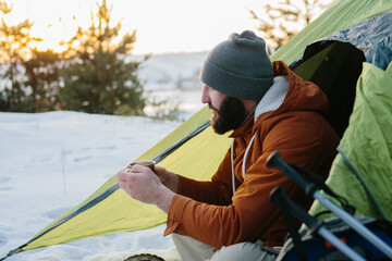 A young bearded man rests in the winter mountains near a tent. A man-traveler with a beard in a cap and a warm jacket warms up by drinking hot tea or coffee after a hike. Travel, lifestyle