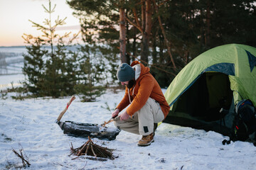 A man sitting near his tent prepares wood wool with a knife for starting a fire in a winter forest at sunset. Winter survival concept.