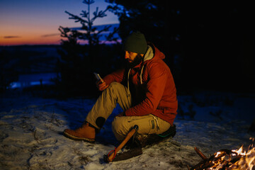 A hiker checks his phone while warming himself by a campfire at night in the mountains.