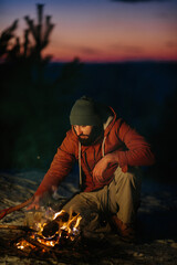 Image of hiker young man cooking dinner by a campfire in the mountains in winter. Male hiker with a beard sitting by the tent after sunset, making a campfire after hiking. Travel, lifestyle concept.