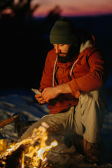 A hiker checks his phone while warming himself by a campfire at night in the mountains.