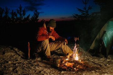 A bearded young man makes delicious coffee on a bonfire while enjoying the sunset in a winter forest. The concept of survival and hiking in the wild.
