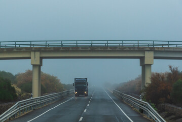 Truck driving on a conventional two-way highway on a foggy day, with the lights on due to poor visibility.