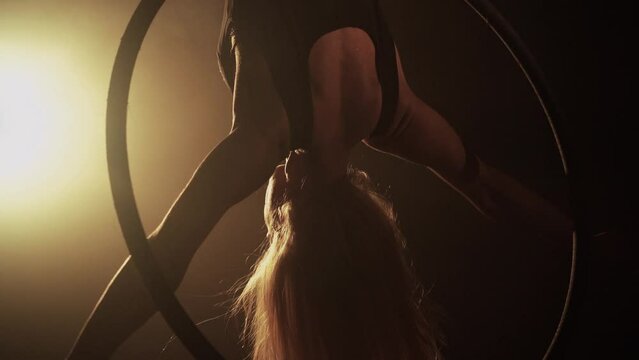 Young woman performs the acrobatic elements in the air hoop. Aerialist in on black background dark studio with backlight. For sports, acrobatic, circus school.