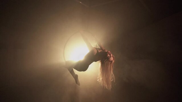 Young woman performs the acrobatic elements in the air hoop. Aerialist in on black background dark studio with backlight. For sports, acrobatic, circus school.