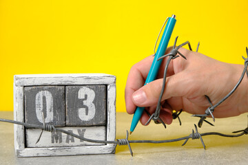 Hand wrapped barbed wire holds ballpoint pen on yellow background.On calendar May 3.Concept for Press Freedom Day.