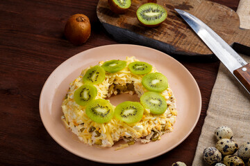 Layered salad with chicken and kiwi next to the table kiwi and quail eggs and a knife.