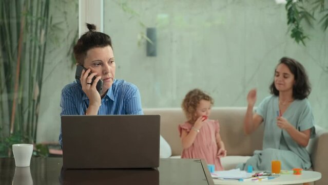 Serious Caucasian Lesbian Woman Making Phone Call While Working On Laptop From Home. Her Wife Playing With Their Daughter On Couch In Background