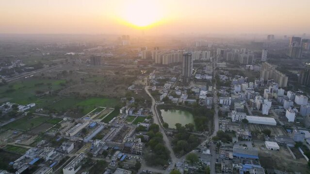 aerial drone shot flying towards sunset with golden red hues showing homes buildings and lakes below and skyscrapers in the distance in the city of gurgaon delhi NCR
