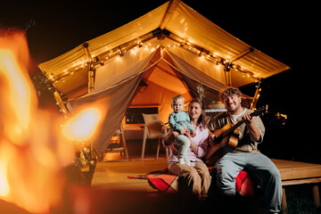 Obraz na płótnie Canvas Happy family relaxing and spend time together in glamping on summer evening and playing guitar near cozy bonfire. Luxury camping tent for outdoor recreation and recreation. Lifestyle concept