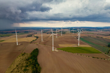 Drone flight at high altitude over a large field with many wind turbines spinning. The wind turbines cast a long shadow because the sun is setting. In the background, at storm sky.