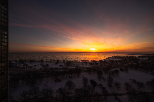 High rise building view of a colorful yellow orange and pink sunrise over Lake Michigan in winter with pieces of ice reflecting the orange glow on the water and snow on the ground between trees.