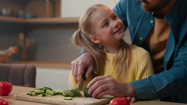 Happy Caucasian bearded father dad teach cute small kid girl child hold knife cut fresh cucumber vegetable for salad in kitchen help little daughter learn cook prepare healthy food cooking at home
