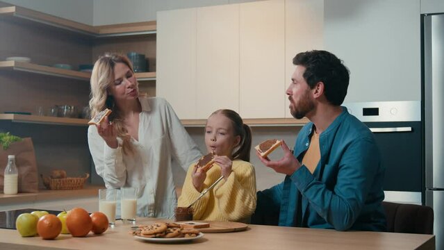 Happy Caucasian family enjoy breakfast together at kitchen. Cheerful parents and cute small kid girl eat bread with chocolate sandwich together dining at table at home. Mother, father and daughter eat