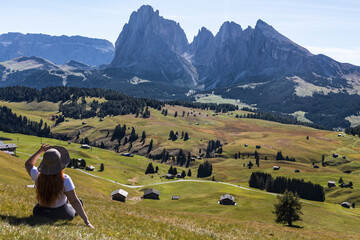 A woman in a straw hat sitting on Alpine meadow admiring the mountain scenery. Alpe di Siusi alpine pasture in South Tyrol, Dolomites, Italy. View of Sassolungo (Langkofel) massif. 