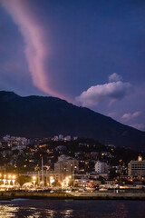 Town On The Coast At Night. A coastal town with buildings and a harbor is nestled at the base of a dark mountain under a dramatic purple and dark blue sky.