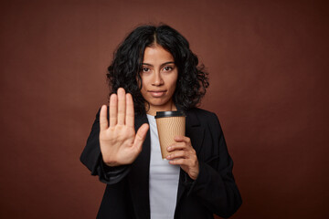 Young business woman drinking a take-away coffee standing with outstretched hand showing stop sign, preventing you. - obrazy, fototapety, plakaty