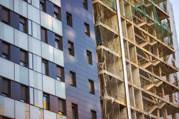 Fototapeta premium Tall high-rise modern apartment or office building facade. Comparison of two houses - unfinished and completed buildings on a construction site. Housing business, real estate investment.