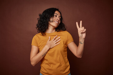Young colombian curly hair woman isolated on brown background taking an oath, putting hand on chest.