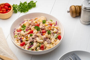 Homemade macaroni salad with elbow pasta and canned tuna, carrot, Purple cabbage, tomato, corn, green peas and mayonnaise dressing in a white plate on wooden table.
