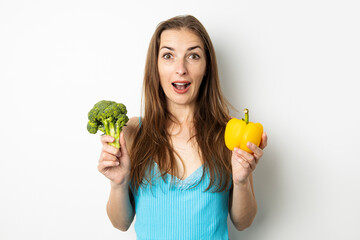 Surprised young woman holding broccoli and paprika on white background