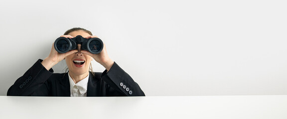 Surprised young woman looking through binoculars peeking out from behind white cardboard. Banner